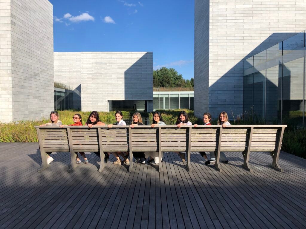 Nine students sit on a long bench and turn around to look at the camera. In the background are the buildings of the Glenstone museum.