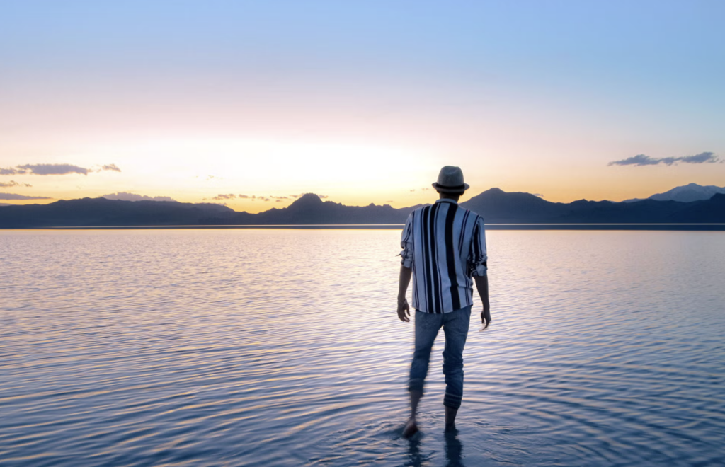 A figure wearing a hat looks out over a sunset over mountains in the distance,  which reflects on the calm water in front of him. 