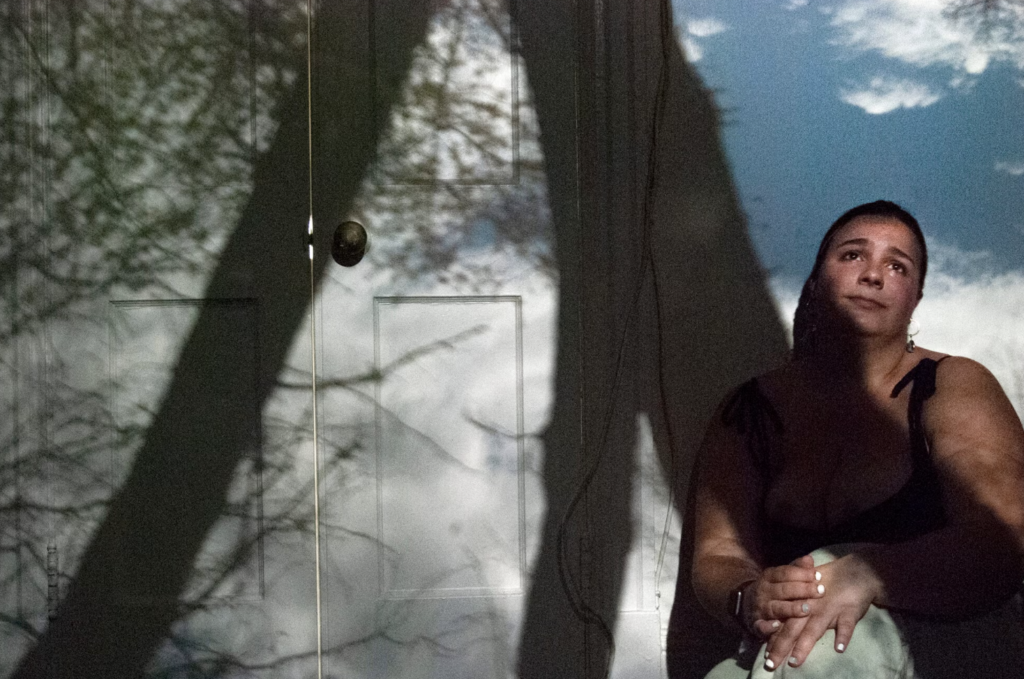 A projection of trees and sky overlayed on a person sitting on the floor. 