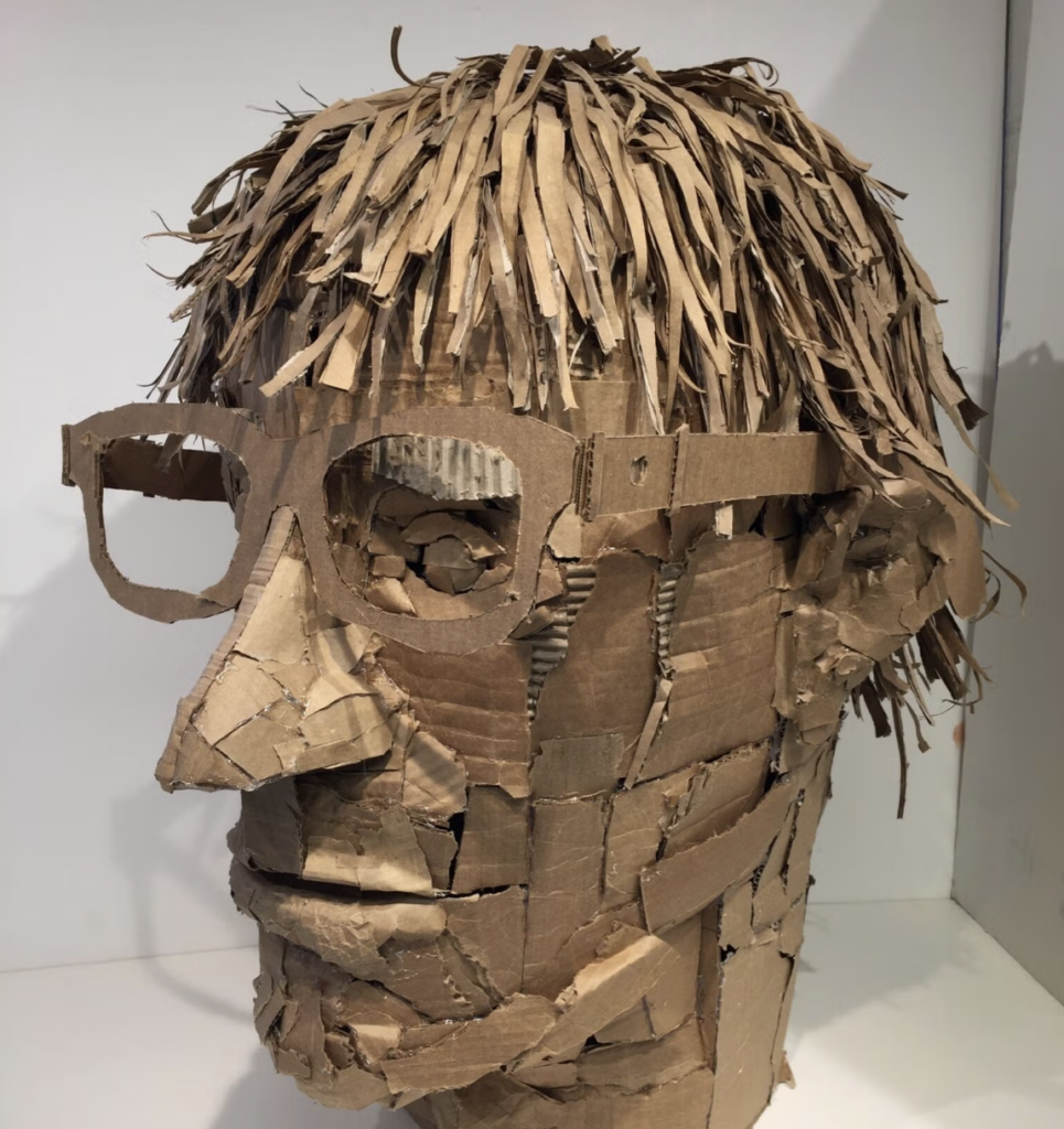 Sculpture of a head with short hair and glasses made out of pieces of cardboard.